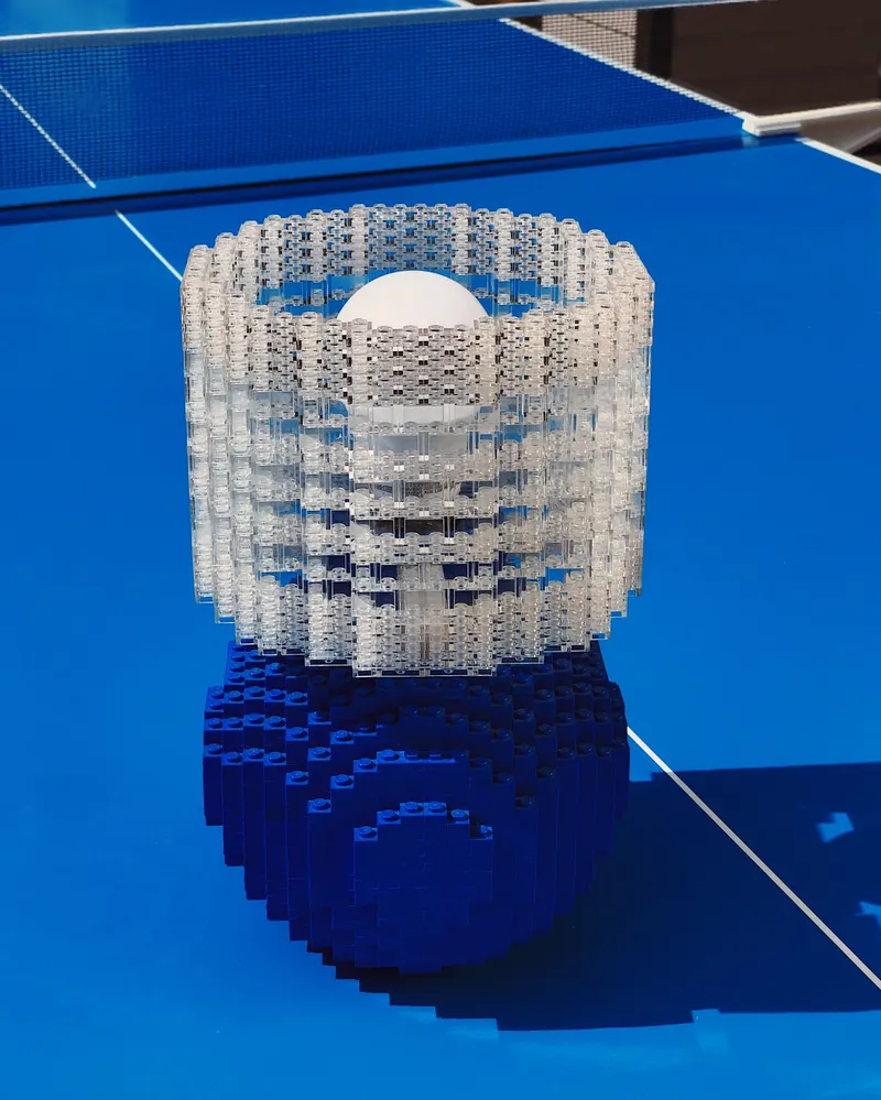 A blue Splash of Color lamp with a clear brick shade lit on a sunny blue ping-pong table outdoors.